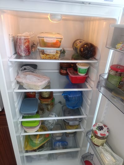 The image shows the interior of a domestic refrigerator. The shelves are stocked with various food items, including jars of preserves, containers of pasta and vegetables, a large plastic bottle of dark liquid, and several smaller food storage containers. The refrigerator appears to be well-used, with some items wrapped in plastic bags. The lighting suggests the refrigerator is in a kitchen environment. No people are visible. The scene is consistent with typical food storage in a residential setting.