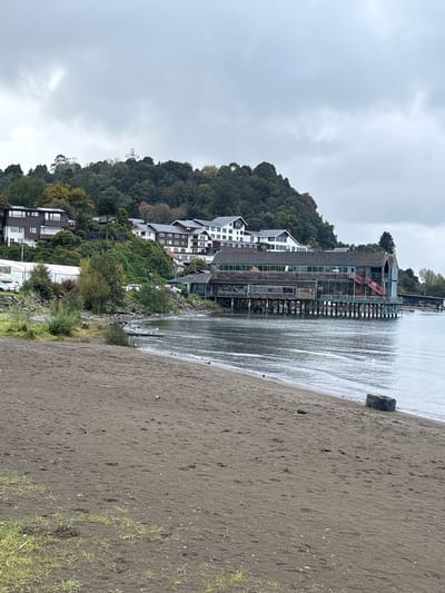 The image captures a view of Puerto Varas, Chile, during overcast weather. The foreground shows a dark sand beach, leading to a body of water, likely a lake. Buildings and a structure on stilts are visible, built into a forested hillside that forms a backdrop. A Christian cross sculpture is visible on the hill. The sky is covered in gray clouds, suggesting an overcast day.