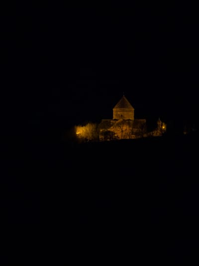 At night, the image shows the lit Kecharis Monastery in Tsakhkadzor, Armenia. The stone structure, including the church with its conical roof, is visible against the dark sky. The lights highlight the building's architecture.