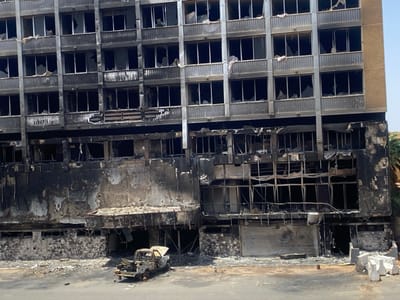 A severely damaged building exhibits extensive fire damage, with blackened walls and shattered windows across multiple floors. A burnt-out vehicle sits in front of the structure. The lower levels are heavily charred, and a partially collapsed entrance canopy is visible. Concrete barriers are scattered nearby, and the entire scene conveys a sense of destruction and abandonment.