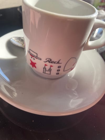 A close-up, eye-level shot captures a white ceramic coffee cup and saucer. The cup features a decorative design with a stylized drawing of a person wearing a hat and scarf, a small castle with flags, and three coffee beans. The word "Reck" is written in cursive script on the cup. A silver spoon rests on the saucer, partially beneath the cup. The saucer and cup are illuminated by a bright light source, casting shadows on the surface below. The background is dark and out of focus, with a hint of a darker surface beneath the saucer. The scene appears to be set on a table in Gerstheim, France.