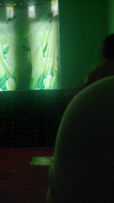 The image shows a person sitting in a room, facing a window. The window has a patterned curtain with light green and white floral designs. The room's walls are painted a dark green color. The person is sitting on what appears to be a bed, with a dark patterned fabric. The bedspread is red. There is a small rectangular object, possibly a book or a piece of paper, lying on the red bedspread. The person is only partially visible in the frame and their face is not visible. The focus of the image is on the person and the patterned curtain. The image was taken inside a room located in the city of Kecamatan Babakan Madang, Indonesia, at the coordinates latitude: -6.561, longitude: 106.863.