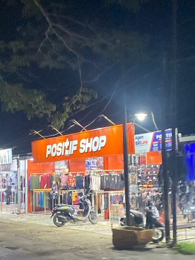 The image depicts a storefront at night, likely in Macege, Indonesia. The primary focus is an illuminated orange sign that reads "POSITIF SHOP" in large white lettering. To the right of the main sign, there are smaller signs, one of which appears to be for "Agen BRIlink". The storefront itself seems to be a market stall or small shop selling clothing, with racks of garments visible behind the counter. A scooter is parked in front of the stall. The background is dark, suggesting it is nighttime, and trees with branches are visible against the dark sky. The overall impression is that of a bustling street-side market operating after dark.