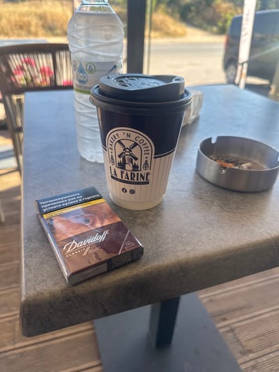 A cigarette pack of Davidoff Classic SL-line is placed on a grey, textured table. Next to it is a black and white paper coffee cup with the "LA FARINE" logo and a picture of a windmill. A plastic bottle of water is visible behind the coffee cup. In the background, an ashtray with cigarette butts and ash is also present. The setting appears to be outdoors, possibly a cafe or restaurant, with a blurred background showing foliage and a road with a vehicle. The scene is well-lit, suggesting daytime.