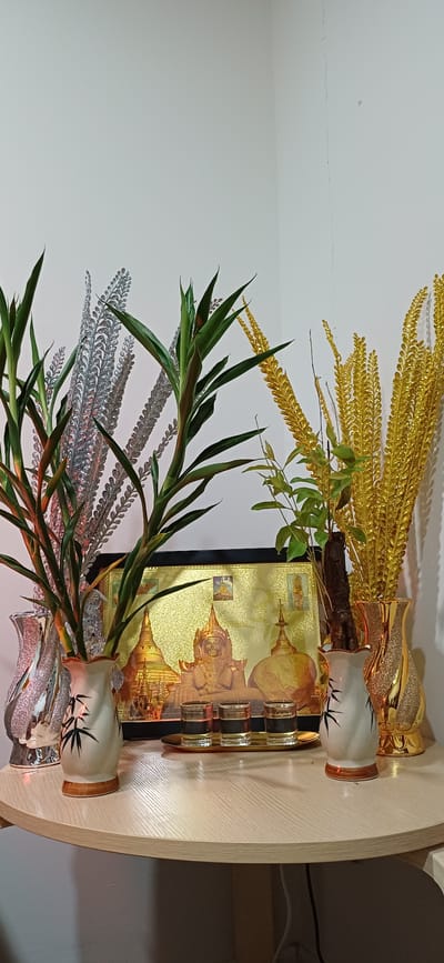 This is a media file of a decorative arrangement placed on a light-colored wooden table. The arrangement features several objects, including two vases, a framed picture, and various decorative branches. One vase is white with black bamboo-like markings and a copper rim, while the other appears to be made of clear, textured glass with glittery elements. A framed image, predominantly gold in color, depicts Buddhist stupas and temples. There are also sprigs of gold and silver decorative foliage, along with green leafy branches, possibly from a plant. A small tray holding what appear to be four small shot glasses with gold rims is visible in front of the framed picture. The overall impression is one of ornate decoration, possibly for a spiritual or celebratory purpose, with the gold elements suggesting prosperity or reverence. The lighting suggests it's indoors, likely during the day, as indicated by the natural light illuminating the scene. The location context provided, Samrong Nuea Subdistrict Municipality, Thailand, suggests a cultural setting where such decorations might be common. No people are visible in the media file, and there is no discernible text.