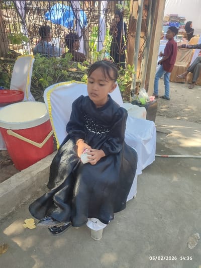 A young girl in a long-sleeved black dress sits on a white chair outdoors. She holds something small and white in her hands. Nearby is a red and white container and a concrete ledge. In the background, people are visible, some near a decorated fence. The date "05/02/2026" and time "14:36" are printed on the bottom right corner of the image.