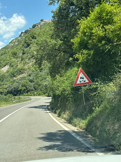 The image depicts a winding asphalt road in Blizikuće, Montenegro, with lush green vegetation on either side and a steep, tree-covered hillside in the background. A white line delineates the lane markings on the road. On the right side of the road, a triangular warning sign with a red border is visible, indicating a slippery road surface, depicted by a car icon with two wavy lines underneath. The sky above is a clear blue with scattered white clouds. A white metal guardrail runs along the left side of the road. In the distance, a few white buildings with red roofs are perched atop the hillside. The scene appears to be captured from within a vehicle, as indicated by a portion of the vehicle's interior visible at the bottom of the frame, casting a shadow on the road.