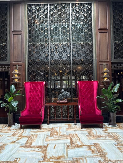 This image depicts an opulent interior space within a building in Tongxiang, China, likely a hotel lobby or reception area. The scene is symmetrically arranged, featuring a seating area against a decorative wall.

Two high-backed armchairs upholstered in deep red velvet, with prominent tufting and brass-colored trim, are positioned facing the viewer. Between these chairs stands a dark brown wooden console table with intricate carved legs. Resting on the table is a dark, irregularly shaped sculptural object, possibly a stylized rock or abstract form, placed on a smaller, ornate wooden base.

Behind this arrangement is a large, multi-panel decorative screen constructed from dark metal or wood, featuring a repeating pattern of overlapping semicircles or fan shapes with subtle metallic accents. This screen is partially transparent, revealing glimpses of an interior space with windows beyond. The decorative screen is flanked and topped by dark wooden wall panels with recessed sections, exhibiting traditional Chinese architectural motifs.

Two wall-mounted sconces, each composed of three tiered, square, amber-toned glass shades, are affixed to the wooden panels on either side of the screen. Large green potted plants, possibly species of ficus or similar broad-leaf plants, stand in dark, decorative planters to the left of the left armchair and to the right of the right armchair.

The floor is covered with large, rectangular, polished marble tiles, displaying a variegated pattern of light cream, beige, and brown striations, laid in a staggered grid. No individuals are present, and no actions or interactions are depicted. The overall environment conveys a sense of traditional luxury and serene grandeur.
