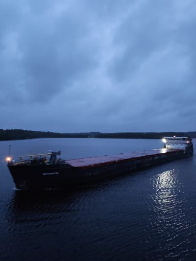 An aerial shot captures a barge moving through the dark waters of Devyatiny, Russia, under a cloudy, twilight sky. The barge, a long, flat vessel with a red cargo bed, is illuminated by lights on its stern and a yellow light on its bow. The boat's name, "BOAT-SMT 136", is visible on its side. The water around the barge reflects the lights, creating shimmering patterns. The background shows a tree-lined shore fading into the distance, with faint lights visible on the horizon.