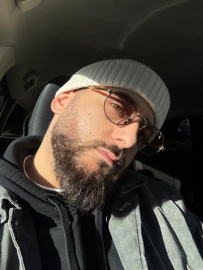 The scene depicts a male individual inside a vehicle in Hérouville-Saint-Clair, France; specific external landmarks are not visible. The subject, positioned centrally, is observed from a slightly low angle. He has a beard and mustache, and is wearing a light grey ribbed knit beanie. Round, gold-rimmed sunglasses with dark lenses are worn, with the right lens flipped upward, revealing a light brown eye. He wears a dark-colored hooded sweatshirt underneath a faded grey or black denim-style jacket. A thin, light-colored necklace is visible. Direct sunlight from the left illuminates the left side of his face and parts of his clothing, creating strong highlights and casting the right side of his face and the vehicle's interior into deep shadow. A dark-colored car seat headrest is visible behind him. No other individuals or significant actions are observed.