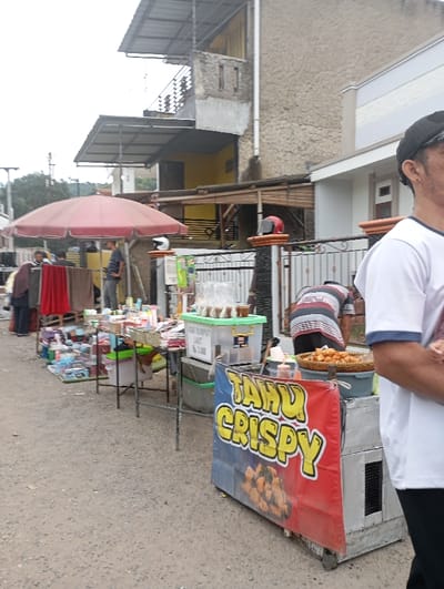 The image presents an outdoor street market scene in Cilampeni, Indonesia, under an overcast sky. The ground is an unpaved dirt surface.

In the right foreground, a male individual, cropped from the chest up, wears a white t-shirt with blue trim and a black cap. He faces away from the camera. Immediately adjacent to him is a mobile vendor cart with a red and blue banner displaying "TAHU CRISPY" in prominent yellow letters, accompanied by an image of crispy fried tofu. On the cart's top surface, a circular woven tray holds a quantity of golden-brown fried food, with condiment bottles positioned nearby.

Behind this cart, a second male individual, viewed from the back, wears a horizontally striped t-shirt (white, red, and black stripes) and dark pants. He is bent over, engaged in activity at the stall.

Further left, a series of informal market stalls are set up. One stall, covered by a large red market umbrella, displays various items of clothing, including red and brown fabrics, hanging on racks. A male figure stands near this clothing stall. Adjacent tables exhibit an assortment of consumer goods, including packaged items and transparent plastic containers. A clear plastic container with a green lid holds cups of liquid, labeled "AGAR RUMPUT LAUT Rp. 3.000" (Seaweed Jelly Rp. 3,000).

The background features residential structures. Directly behind the food cart stands a two-story building with a light-colored lower façade and a textured, light brown upper section. Its ornate dark metal gate has a red motorcycle helmet resting on one of its posts. To the left, a multi-story concrete building, appearing unfinished or unrendered, features multiple corrugated metal awnings extending from its upper levels. Sections of its interior walls are painted yellow. Distant hills with green foliage and utility poles are visible in the far left background.