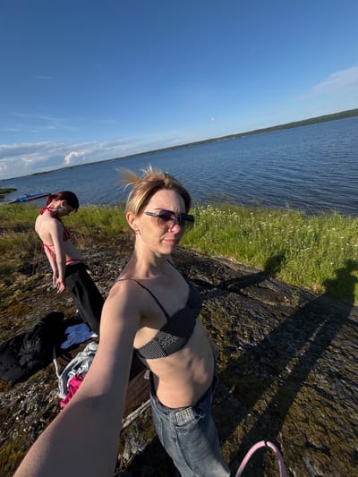 Two women are standing on a rocky shore by a large body of water under a bright blue sky with some clouds. The woman in the foreground is taking a selfie, wearing a black and white polka dot bikini top and blue jeans. She has blonde hair pulled back and is wearing sunglasses. Behind her, the second woman is seen from the back, wearing a red and white striped bikini top and black pants. She is looking down, with her hands behind her back. The environment is natural, with grass and small white flowers growing on the rocky terrain. In the distance, the water stretches out, with a tree-lined shore visible on the horizon. There are also some boats docked near the shore in the background. The scene suggests an outdoor recreational activity in a natural setting, likely during daylight hours. The specific location is identified as Shuiskaya Chupa, Russia.