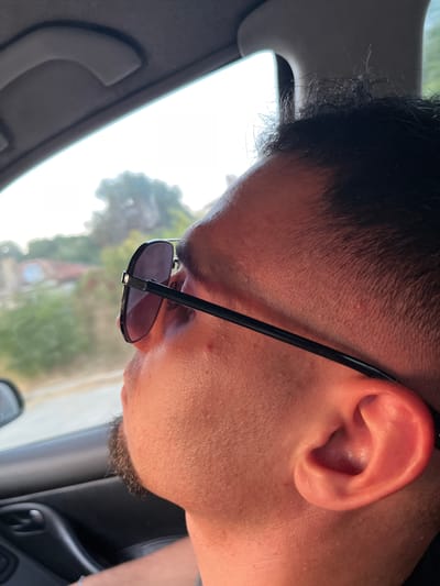 The image is a close-up, side profile of a person wearing sunglasses, seen from a slightly high angle. The person's right ear is prominently visible, with skin showing texture and a slight pinkish hue. Their hair is dark and short, with some lighter, finer hairs visible near the temple. The sunglasses have black frames and dark lenses, obscuring the eyes. The lower part of the person's face, including the chin and a small patch of beard stubble, is also visible.
The setting appears to be inside a vehicle, with a portion of the car's interior, including a gray handle and part of the roof lining, visible in the foreground. Through the window, a bright, overexposed sky is visible, suggesting it is daytime, possibly midday, with strong sunlight. In the background, out of focus, there are hints of greenery, suggesting an outdoor environment. A building with a reddish roof is also subtly visible. The overall lighting suggests a bright, possibly sunny day. No text is visible in the image. The image is oriented sideways.