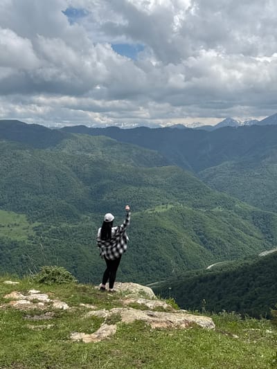 A person stands on the edge of a rocky cliff overlooking a vast, green mountain landscape. The person, viewed from behind, is wearing a white baseball cap, a black and white checkered shirt over a white t-shirt, black pants, and dark shoes. Their right arm is raised, and their fist is clenched. The mountains are covered in dense green forest, with layers of ridges receding into the distance. The sky is cloudy, with patches of blue visible. In the far distance, snow-capped peaks are visible. The foreground consists of grass and rocks. The location is identified as Nizhnyaya Teberda, Russia.