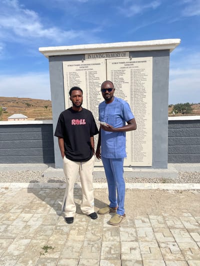 The scene takes place in Zot Shen, Nigeria, where two adult males stand in front of a concrete memorial structure. The male on the left, a younger adult with a dark beard and short dark hair, wears a black short-sleeved t-shirt with "ROLL" in red lettering, light beige loose-fitting trousers with dark vertical accents and a circular patch on the left leg, and black slip-on shoes. His hands are in his pockets. The male on the right, an older adult with a clean-shaven head and dark sunglasses, is dressed in a light blue traditional Nigerian tunic and matching trousers, with light brown slip-on sandals. He holds a white smartphone in his left hand. Both individuals face the camera, posing side-by-side.

The memorial behind them features a light gray base and a lighter top slab. Its front face contains dark rectangular plaques listing names under the headings "IN LOVING MEMORY OF" and "VICTIMS OF 7TH MARCH, 2010 MASSACRE AT DOOD NAHAWA, PLATEAU STATE." The ground is paved with light-colored interlocking stones, with some sparse vegetation growing through the cracks. The background reveals distant dry, undulating hills and a clear blue sky with scattered white clouds. A light-colored building with a red roof is visible on a hill in the far left background. Bright sunlight illuminates the scene, casting shadows to the right and slightly behind the subjects.