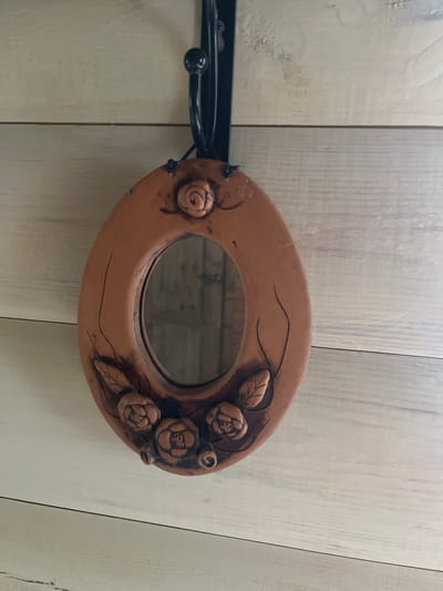 An oval, terra cotta mirror with floral embellishments hangs on a black metal hook against a light-colored wooden plank wall. The mirror is positioned in the center of the frame, slightly tilted. The wooden wall has a textured, slightly aged appearance. The image does not contain any people or show any specific location within Староживотинное, Russia.