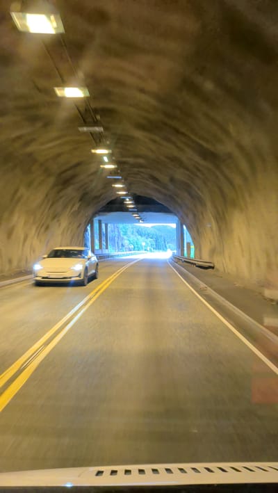 A white electric car drives through a tunnel in Mosjøen, Norway. The tunnel is made of rough-hewn rock and lit by rows of fluorescent lights. The road is paved and marked with yellow double lines. Outside the tunnel, the sky is bright, and trees and mountains are visible. The car is the only vehicle in the tunnel, and no people are visible. The scene is captured from the perspective of another vehicle, looking forward.