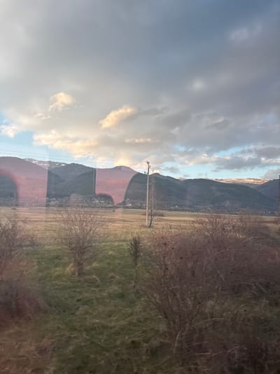 This image depicts a rural landscape near the city of Lesnovo, Bulgaria, viewed through a reflective surface, likely a window. The scene is dominated by a range of mountains in the background, their upper slopes covered with patches of snow. At the base of these mountains, a scattered village or settlement with numerous small buildings is visible. The midground features a wide, open field with sparse, dry brown and green grass. A single wooden utility pole stands prominently in the field, slightly to the left of the frame's center. Overhead power lines traverse the scene horizontally, connecting to this pole and a partial view of a metal lattice power transmission tower on the far right. The foreground is blurred and occupied by dense, leafless shrubbery and bare tree branches. The sky is largely overcast with significant grey cloud cover, interspersed with patches of blue sky and areas where sunlight illuminates the clouds, suggesting either late afternoon or early morning. Prominent, distorted reddish and lighter reflections on the viewing surface partially obscure the mountain range and the sky. No individuals or animals are visible.