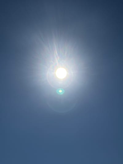 The image is a photograph of the sun in a clear blue sky. The sun is bright and appears in the upper center of the image, with visible sun flares radiating outwards. There are lens flares present, one large and circular, and a smaller, greenish circular flare below the sun. The sky is a uniform, deep blue, indicating it is daytime with no clouds. There are no people, buildings, or any other objects visible in the scene. The location in Oława, Poland, is not discernible from the image itself, as it depicts a universal celestial phenomenon.