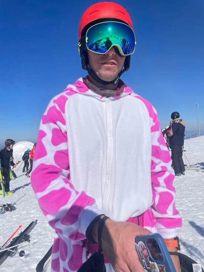 The image captures a scene on a snow-covered mountain slope in Jahorina, Bosnia and Herzegovina, under a clear blue sky. The primary subject is an adult male positioned centrally, facing the camera. He wears a red ski helmet, reflective green-blue ski goggles that mirror the surrounding snow and sky, and a white and pink cow-print hooded one-piece ski suit. A patterned neck gaiter is visible beneath his chin. His right hand, extended slightly forward, holds a blue smartphone decorated with stickers. In the background, multiple other individuals are visible on the snowy terrain, equipped with ski gear including helmets, jackets, skis, and poles. Some appear to be standing or conversing. Ski equipment, including several pairs of skis and poles, is lying on the snow near the foreground subject and in the mid-ground. A ski lift support structure is partially visible in the distant background on the right side of the frame.