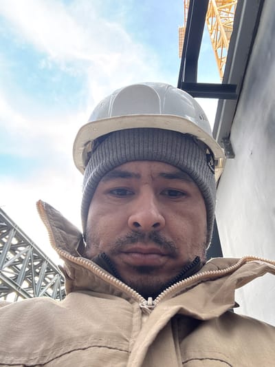 A man wearing a white hard hat and gray beanie looks directly at the camera. He is also wearing a tan jacket with the zipper slightly open. Behind him is a steel construction structure and a glimpse of a crane. The sky is visible with light clouds, suggesting an outdoor environment. The image has an unusual perspective, taken from a low angle and rotated.