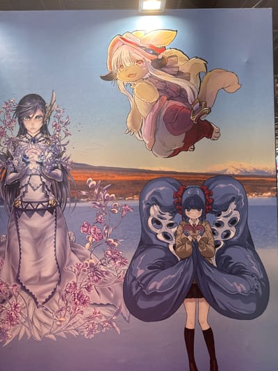 An anime-style artwork is displayed on a large, flat surface, possibly a wall or banner, located at the Parc des Expositions in Villepinte, France. The artwork features three distinct characters against a backdrop of a serene landscape with a body of water and distant, snow-capped mountains.
In the upper portion of the artwork, a fluffy, light-colored creature with large ears and yellow eyes floats in the air. It is wearing a red garment and a white and pink layered skirt.
To the left, a female character with long, dark hair, green eyes, and wearing an intricate white gown adorned with geometric patterns and gold accents, stands with her hands held together. She is surrounded by delicate pink and purple flowers and small, sparkling droplets.
In the lower center, a young female character with dark hair, red ribbons in her hair, and large, dark eyes stands. She is wearing a brown jacket over a white shirt and a dark pleated skirt, with dark socks and shoes. She is holding a heart-shaped object. Enclosing her are two large, dark blue, bulbous forms with multiple eye-like structures, giving the impression of a protective embrace or a symbiotic entity.
The overall lighting suggests an indoor environment with a bright light source visible at the top, possibly a projector or ceiling light, casting a subtle glow on the upper edge of the artwork.