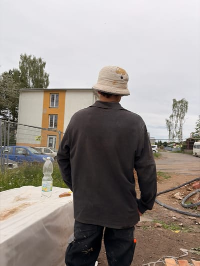 A person wearing a tan bucket hat and a dark long-sleeved shirt stands with their back to the camera at a construction site in Příbram, Czechia. The person is wearing dark pants with grey accents, splattered with what appears to be mud or dust. In the foreground, a white sheet covers a surface, upon which sits a plastic bottle of "DOBRÁ VODA" and a small, light brown rectangular object, possibly a piece of brick or cake. Behind the person, a multi-story building with white and orange sections is visible, with several windows. A chain-link fence encloses the construction area, with a blue car and other vehicles parked beyond it. A large tree with sparse foliage stands to the right, and a cloudy sky completes the scene. The ground is dirt and debris, with construction materials like hoses and bricks scattered around.