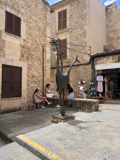 In the town square of Pollença, Spain, a group of people are seated on a stone bench next to a contemporary sculpture of an abstract bird. The sculpture is dark bronze, with elongated limbs and a slender body, casting a distinct shadow on the cobblestone pavement. To the left, two young women sit on small pink stools near a window with brown shutters. One woman wears a pink tank top and denim shorts, while the other has on a white dress. Across from them, further down the bench, another woman in denim shorts and a black top is seated. On the right side of the square, a woman wearing overalls stands near a shop that sells clothing and accessories. She is holding a drink in her hand. The buildings surrounding the square are made of rough-hewn stone, with wooden shutters on the windows. The ground is paved with irregular cobblestones. The sky is a clear blue with some light clouds.