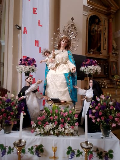The image shows a religious display, likely in a church or religious setting in Municipio Tarija, Bolivia.

At the center is a statue of the Virgin Mary holding the baby Jesus. Mary is wearing a white dress with a blue and gold cloak. She has a crown-like halo with star-shaped spikes around her head. The baby Jesus is also wearing a light-colored dress.

On either side of the Virgin Mary are statues of religious figures, likely saints, in white and black robes with their arms outstretched towards Mary.

The statues are placed on a table covered with a white cloth. On the table are floral arrangements of pink, white, and purple flowers, as well as candlesticks with white candles. The cloth is decorated with a design of grapes, leaves, and a chalice.

In the background, there's a banner hanging with red letters and what appears to be another religious statue within an alcove. The interior architecture suggests a church or religious building.