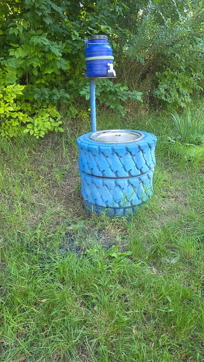 A makeshift water dispenser is situated outdoors in a grassy area with a dense green foliage background. The dispenser consists of a blue plastic barrel with a black lid, mounted on a blue metal pole. A white faucet protrudes from the side of the barrel. Below the barrel, two stacked, blue-painted tires form a base. A metal basin with a drain is inserted into the top tire. The area around the dispenser is covered in green grass with some scattered leaves and debris. This scene is located in the village of Grătiești, Moldova.