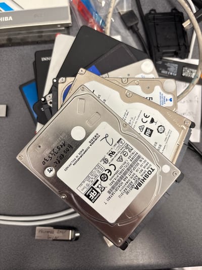 The image shows a pile of computer hard drives and other electronic components on a dark gray surface. The hard drives are of various brands, including Toshiba. One of the hard drives has handwritten labels on it. In the foreground, a small USB drive with a handwritten label is visible. The background shows some cables and what appears to be a black computer accessory. The scene is consistent with a workspace or a collection of electronic devices. There is no visible indication of the city of Mülheim an der Ruhr, Germany, in the image.
