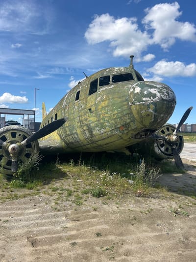 An abandoned, weathered airplane with peeling green paint sits on a gravelly, grassy lot under a partly cloudy blue sky. The plane's windows are broken or missing, and its propellers are intact but appear rusted. In the background, a building with a sign reading "4" and a red structure with water spraying from it are visible. Distant buildings and trees can also be seen on the horizon. The scene appears to be in an industrial or rural area. No people are present.