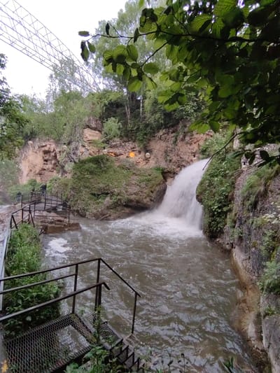 The image depicts a natural scene with a waterfall cascading into a turbulent river. The waterfall is situated amidst rocky cliffs covered with lush green vegetation. A metal staircase with handrails leads down towards the water, suggesting a pathway for visitors. Several people are visible on the walkway near the top of the stairs, observing the waterfall. Overhead, string lights with round bulbs are suspended, indicating a potentially recreational or tourist area. In the background, a large, white, lattice-like structure is visible against the sky, possibly a bridge or part of an attraction. The overall environment is natural and somewhat wild, with water, rocks, and abundant greenery dominating the frame. The city of Красный Курган, Russia is mentioned, but its specific relation to this exact location is not visually confirmed within the image itself.
