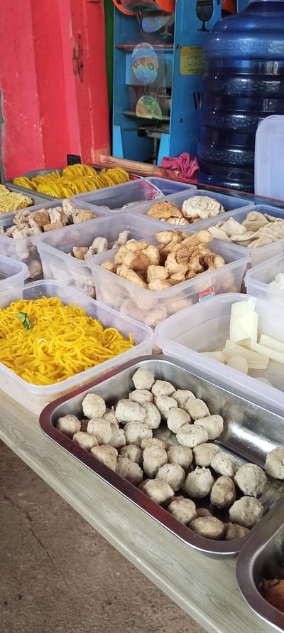 A table in Cimaung, Indonesia (latitude: -7.074, longitude: 107.547) is set up with multiple clear plastic containers filled with various food items. The containers are organized in a row, starting from the left, with yellow noodles, then fried meat chunks, followed by larger pieces of fried meat, and finally, white pieces of unknown food. At the end of the row, there is a large, stainless-steel container with a generous amount of round white meat balls. The table is made of wood, and the background includes a red wall and a blue machine with a water dispenser. The food items are arranged neatly, and the overall scene is clean and well-lit.