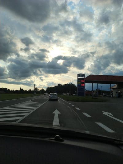 The image shows a view from a vehicle driving on a road in Bernartice, Czechia. In the foreground, the dashboard and windshield are visible. The road has white lane markings and a pedestrian crossing. To the right, a gas station with a price sign displaying "FATO," "DIESEL 37.60," "NATURAL 37.60," and "LPG 13.30" is present. The sky is partly cloudy with sunlight breaking through. Several cars are visible on the road, and trees line the background.