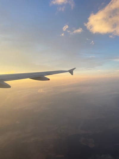 The image is taken from an airplane window, showing an airplane wing extending into the frame from the left. Below the wing, a layer of clouds is visible, obscuring the ground. The sky above is a gradient of blue and yellow, with scattered clouds, suggesting it is either sunrise or sunset. The ground below the clouds appears dark and indistinct, with patches of lighter brown and gray, possibly indicating agricultural fields or a sparsely populated area. There are no people or specific landmarks visible in the image that would allow for precise identification of the location.