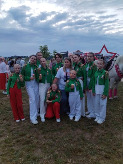 A group of thirteen people, primarily young females, are posing for a photograph outdoors. The individuals are dressed in matching green jackets with ornate gold epaulets and red or white pants. Several of the females are holding ice cream cones. The background suggests a public event, with a white horse and a large red star decoration visible. The sky is overcast with dramatic clouds. The ground is covered in grass, indicating an outdoor, possibly rural setting. The location is identified as Буйніцкі сельскі Савет, Belarus.