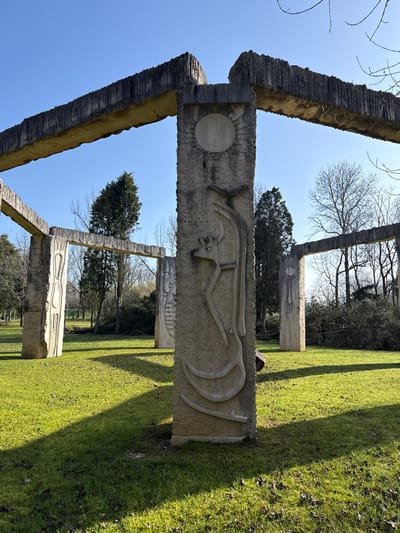The image displays a multi-component architectural sculpture composed of textured concrete or stone elements, located in a grassy park area in Mealhada, Portugal. A prominent vertical column stands in the center foreground, featuring carved bas-relief details including a circular disc at its apex and abstract, elongated forms below. This column is connected to thick horizontal beams extending overhead to the left and right, exhibiting a coarse, striated texture.
Further into the scene, additional vertical columns are visible, forming archways or frames. These columns also bear abstract carvings. The ground is covered with bright green grass, across which long, distinct shadows are cast from left to right by the structures, indicating direct sunlight. The background features a mix of vegetation, including dark green evergreen trees and deciduous trees with bare branches. A clear, light blue sky occupies the upper portion of the frame. No individuals are present in the scene.