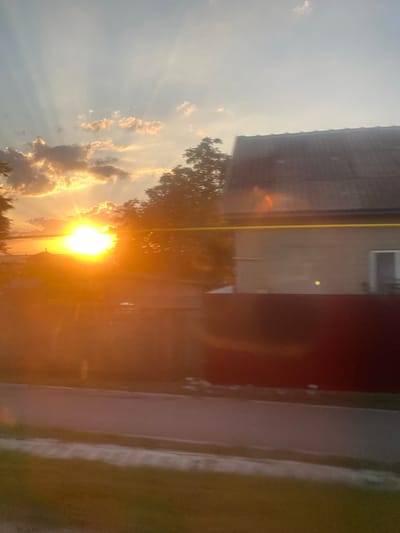 The image depicts a sunset over a rural area. The sun is a bright orb in the sky, casting golden rays through the clouds. Silhouetted against the bright light are trees, houses, and a fence. A yellow line is visible horizontally across the frame, potentially a wire or a painted marking. The foreground shows a road and a grassy area. The overall impression is one of a peaceful evening in a village. The mention of Balabanu, Moldova suggests a specific location within a rural context, likely characterized by similar scenery.