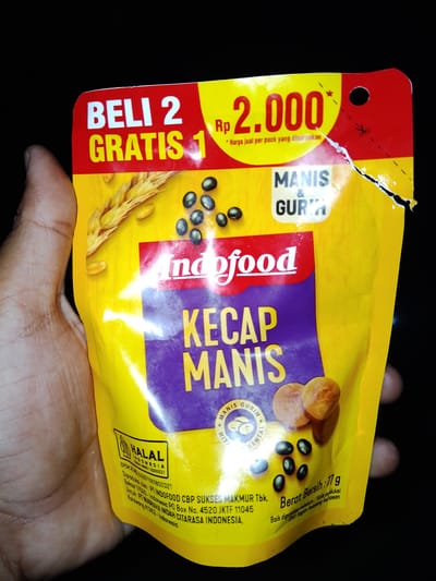 The image shows a yellow pouch of Indofood Kecap Manis (sweet soy sauce) being held by a person's hand. The pouch has promotional text "BELI 2 GRATIS 1" (Buy 2 Get 1 Free) and a price of "Rp 2.000". It also features images of wheat grains and soybeans. The brand name "Indofood" is prominently displayed. There are also claims of it being "MANIS & GURUH" (sweet and savory). Below this, there is a "HALAL INDONESIA" certification. Further text indicates production by "PT INDOFOOD CBP SUKSES MAKMUR Tbk." in Bekasi, Indonesia, and for "PT NUGRAHA INDAH CITARASA INDONESIA" in Karawang, Indonesia. The net weight is stated as "77 g". The image does not contain any information or visual cues related to the city of Sendangrejo, Indonesia.