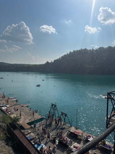 This is a high-angle, outdoor, daytime photograph of a quarry lake in Karyer Izvestnyak, Russia, on a sunny day with scattered clouds. The lake's water is a vibrant turquoise. The foreground features a wooden dock with people sunbathing, swimming, and engaging in recreational activities. Several individuals are visible on the dock, some lying on towels, others standing or walking. A small group of people are in the water near the dock. In the middle ground, various watercraft, including paddleboats and small rafts, are scattered across the lake. The background is dominated by a tree-covered hillside that forms the perimeter of the quarry. Sunlight glints off the surface of the water, creating a shimmering effect. There are structures on the dock that appear to be for recreation, including a swing-like apparatus.