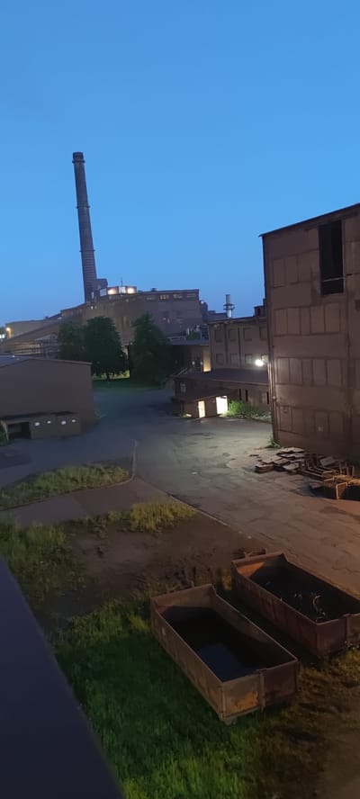 The image depicts an industrial area at dusk. In the background, a tall, slender industrial chimney pierces the clear blue sky. The chimney is part of a larger complex of industrial buildings, some of which are illuminated with warm light from windows, suggesting internal activity. In the foreground, there are several large, open-top industrial bins or dumpsters, some appearing empty and others containing dark materials, possibly waste or debris. The ground is a mix of paved areas and patches of overgrown grass. The scene conveys a sense of industrial activity during twilight hours, likely in an area of Salzgitter, Germany, known for its industrial heritage, particularly in steel production.
