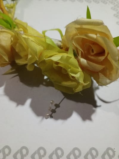 Three artificial, pale yellow roses are arranged closely together. A strand of small, translucent beads extends from the central rose. Green stems and leaves are visible between the flowers. The arrangement casts a dark shadow on a white surface with a faint, decorative pattern along the bottom edge. The roses appear to be made of fabric, possibly silk or satin.