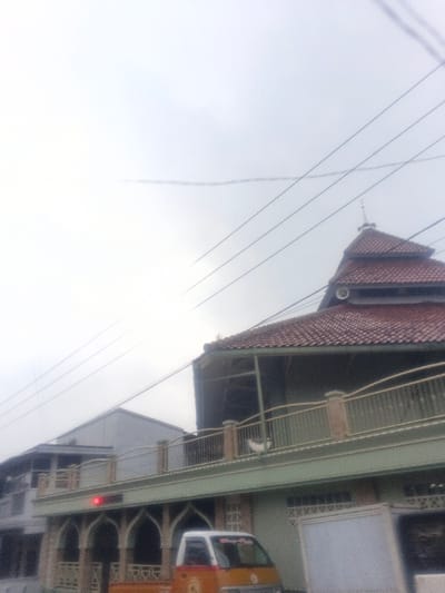 The image depicts an overcast daytime scene in the city of Dakshin Hichachhara, India. The primary subject is a multi-story building, likely a mosque, dominating the right side of the frame. This building features a distinctive multi-tiered, red-tiled roof, topped with a small, pointed finial. A white horn loudspeaker is mounted on an upper section of the roof. Below the roofline, an open balcony or veranda with a green railing and light brown pillars is visible, where a small white object rests on the floor. The lower facade of this building is light green/teal, characterized by arched entryways or windows with decorative latticework. A red light or digital display is embedded in this lower facade. To the left and slightly behind, a second, simpler multi-story building with a light gray facade and a flat or low-pitch roof is partially visible. Multiple dark utility lines traverse the overcast, bright white sky. In the foreground, positioned in front of the main building's lower level, is a yellow and orange small utility truck or pick-up vehicle. No people are discernible in the scene.