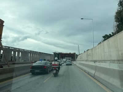 The image shows a view from inside a vehicle traveling on a multi-lane road in Polichni Municipal Unit, Greece. The road is flanked by concrete barriers. On the left, there is a large construction site with unfinished concrete structures. On the right, a concrete wall rises, with some trees and vegetation visible at the top. Several vehicles are on the road, including a black Volvo sedan with license plate BG 3078 HA, a scooter with a delivery box on the back, and other cars. The sky is overcast with heavy clouds. The scene appears to be a section of highway or a major road undergoing construction.