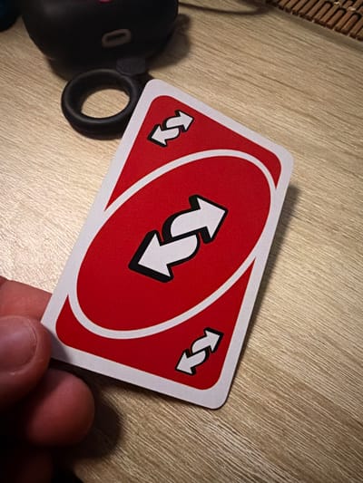 An Uno reverse card, with its iconic red backing and white arrows indicating a change in direction, is held by a hand. The card is positioned diagonally on a light-colored wooden surface, likely a table. In the background, out of focus, a black, circular object with a ring attached is visible, along with a portion of a woven matting material. The lighting suggests an indoor environment, possibly a home. The location of Castillo Bajo-conejito, Spain, is not depicted in the image and cannot be determined from the visual information provided.