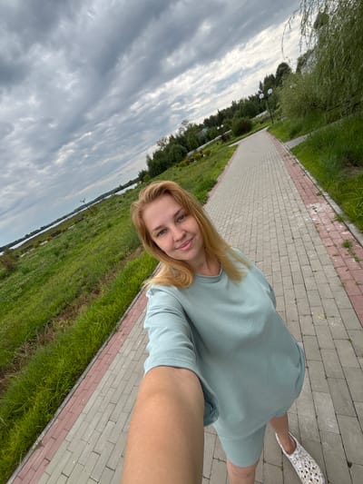 A medium-shot selfie shows a young blonde woman with freckles posing on a paved path beside a grassy embankment. She is wearing a light blue loungewear set and white patterned slip-on shoes. The path is made of interlocking rectangular pavers in shades of gray and pink. The embankment is covered in lush green grass and scattered wildflowers, sloping down towards a body of water visible in the background. A line of trees borders the far side of the water, and the sky above is filled with dramatic, cloudy formations. The scene appears to be in a park or recreational area. This is likely the embankment in Breytovo, Russia, along the Rybinsk Reservoir, a popular location for walks and outdoor activities.