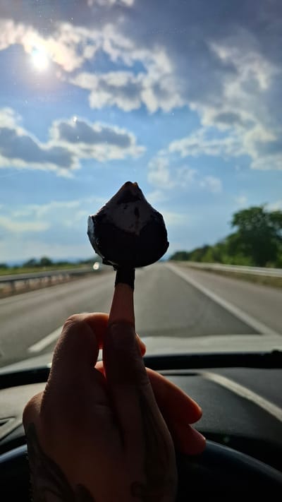 A hand holds a dark, irregularly shaped object, possibly a chocolate-covered fig, on a finger against a backdrop of a highway and a cloudy sky. The sun is visible in the upper left corner, casting a lens flare. The scene appears to be from inside a vehicle, suggested by the presence of a steering wheel and dashboard. The highway stretches into the distance, bordered by trees and a guardrail. The image is taken from a first-person perspective, as if the viewer is the one holding the object. There are no other people visible in the frame, and the location is identified as Shtarkovo, Bulgaria.