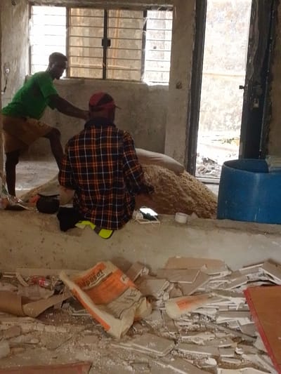 Inside a structure in Agukwu Nri, Nigeria, two adult males are present in an unfinished or disused concrete room with debris on the floor. To the left, a dark-skinned male wearing a green short-sleeved shirt and light brown shorts stands with his body angled, leaning forward, facing right towards the center. In the center, a dark-skinned male wearing a red and black plaid long-sleeved shirt and a red baseball cap sits or kneels on a raised concrete ledge, facing away from the viewer towards a large mound of light brown granular material (possibly sawdust or sand) behind him. A black object, a white smartphone with a charging cable, and a roll of white tape are visible on the ledge near the seated male. The floor in the foreground is covered with broken light-colored tiles, a red plastic bottle, and a crumpled white and orange "Supaset" bag. The room features a window with metal bars on the left wall and an open doorway on the right, revealing an exterior area with dirt and debris. A blue plastic barrel or container is positioned near the doorway.