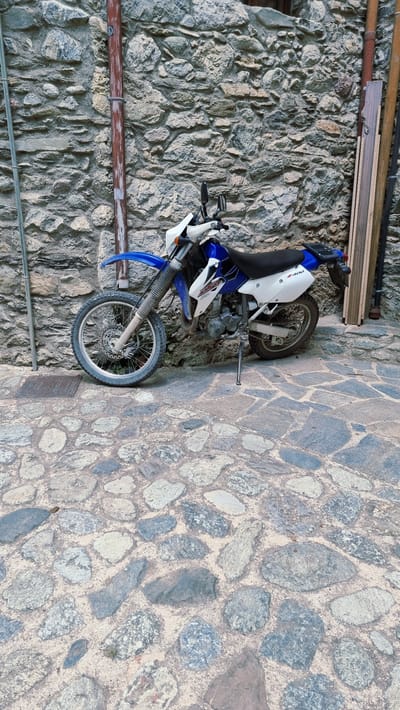 A blue and white dirt bike is parked on a cobblestone street in front of a rough stone wall. The motorcycle, a Suzuki DR-Z400, is upright, supported by its kickstand. It has a black seat, silver handlebars with mirrors, and knobby tires suitable for off-road use. A brown pipe runs vertically along the stone wall to the left of the motorcycle. To the right of the motorcycle, leaning against the wall, are several pieces of wood. The cobblestone street is made up of irregularly shaped grey and tan stones. The scene appears to be in an outdoor alley or street in Camprodon, Spain.
